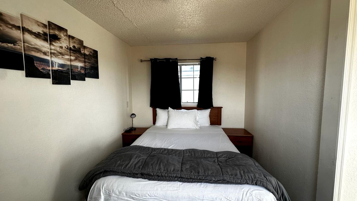 A single bed with a gray blanket, white pillows, and a wooden headboard. There is a window with black curtains, a bedside table with a lamp, and a wall-mounted picture frame.