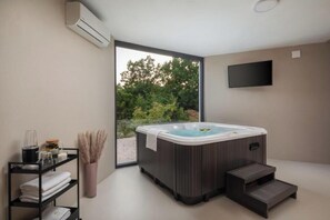 Ein modernes Badezimmer mit Whirlpool, einer Ablage mit Handtüchern, einem Fernseher und Blick auf Bäume.