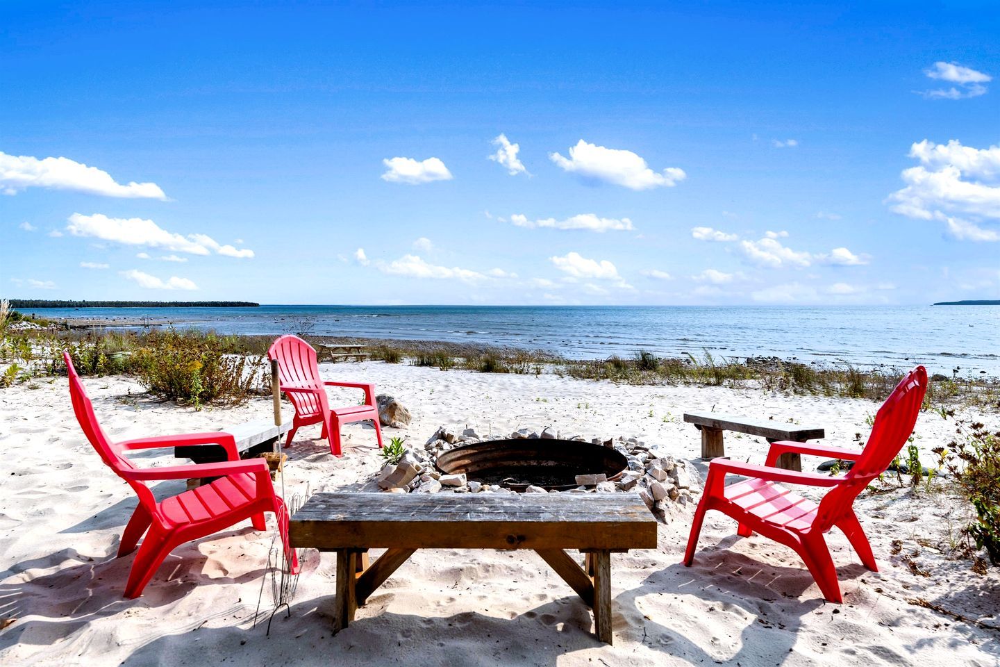A beach setting with red Adirondack chairs, a wooden table, and a fire pit.