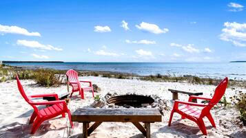 A beach setting with red Adirondack chairs, a wooden table, and a fire pit.