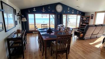 Un coin repas avec une table et des chaises en bois, un ventilateur de plafond et une vue sur la plage grâce à de grandes fenêtres.