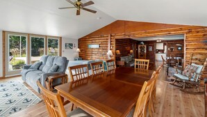 A spacious living area with a large wooden dining table, a grey sofa, and a ceiling fan.