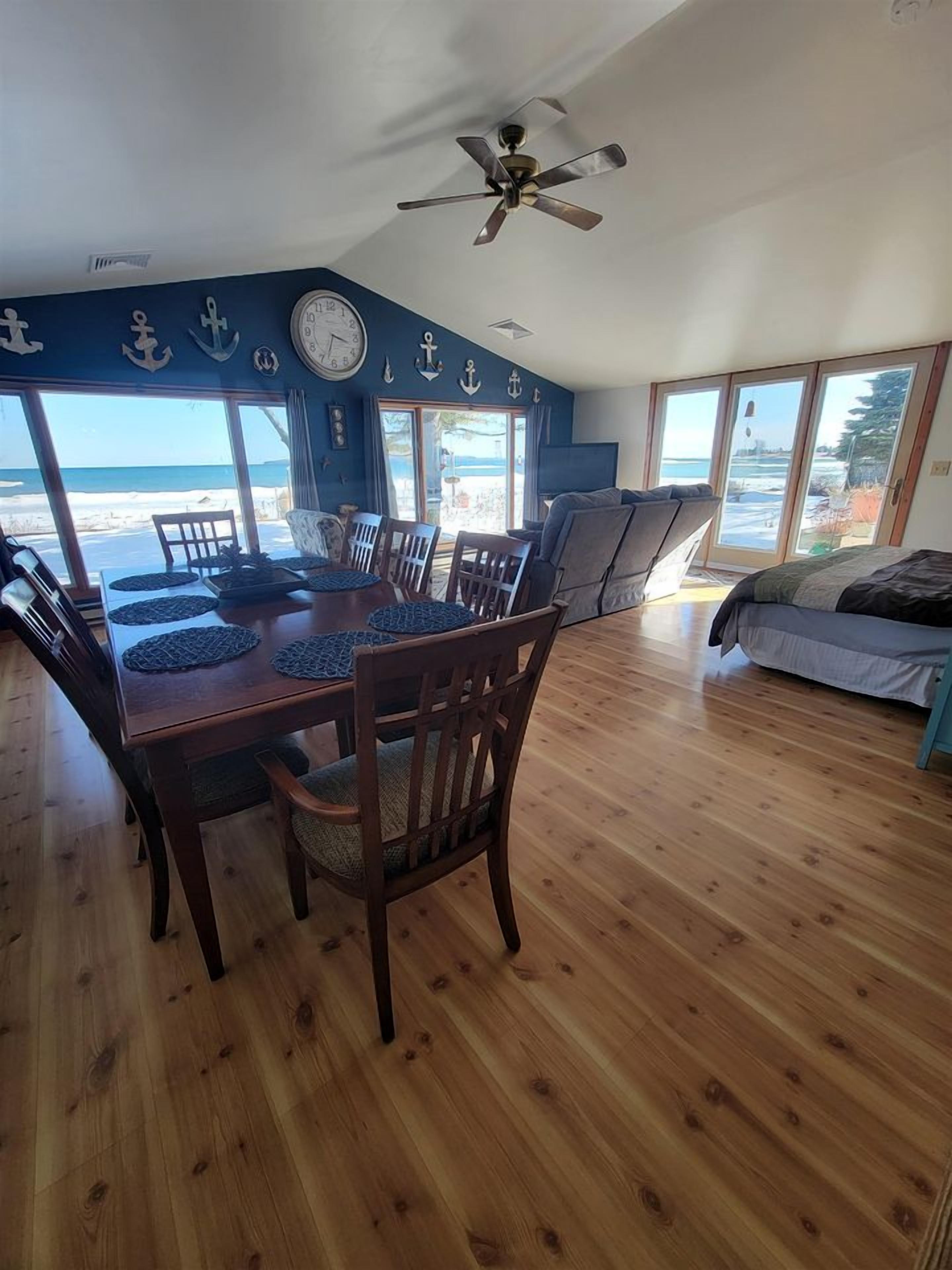 Un comedor con una mesa y sillas de madera, un ventilador de techo y ventanas grandes con vista a la playa.