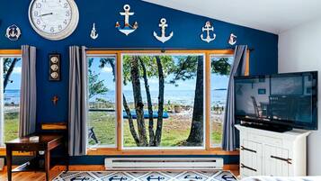 A room with a large window overlooking a beach, a clock on the wall, and a television on a cabinet.