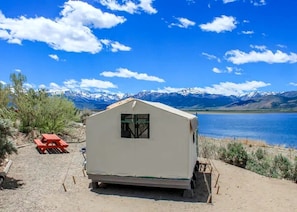 Una caravana de una habitación con ventana, una mesa de picnic y vistas a montañas y un lago.