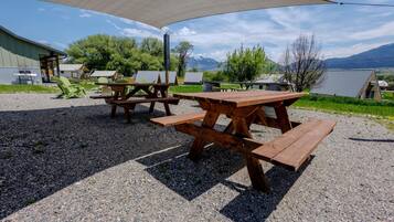 Outdoor picnic tables under a shade structure, with a gravel ground and buildings in the background.