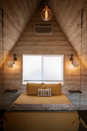 A bedroom with a sloped ceiling, a large window, a bed with pillows, and wall-mounted lighting. - Charming A-Frame Cabins with Shared Kitchen, BBQ & Mountain Views in Emigrant, Montana (Emigrant)