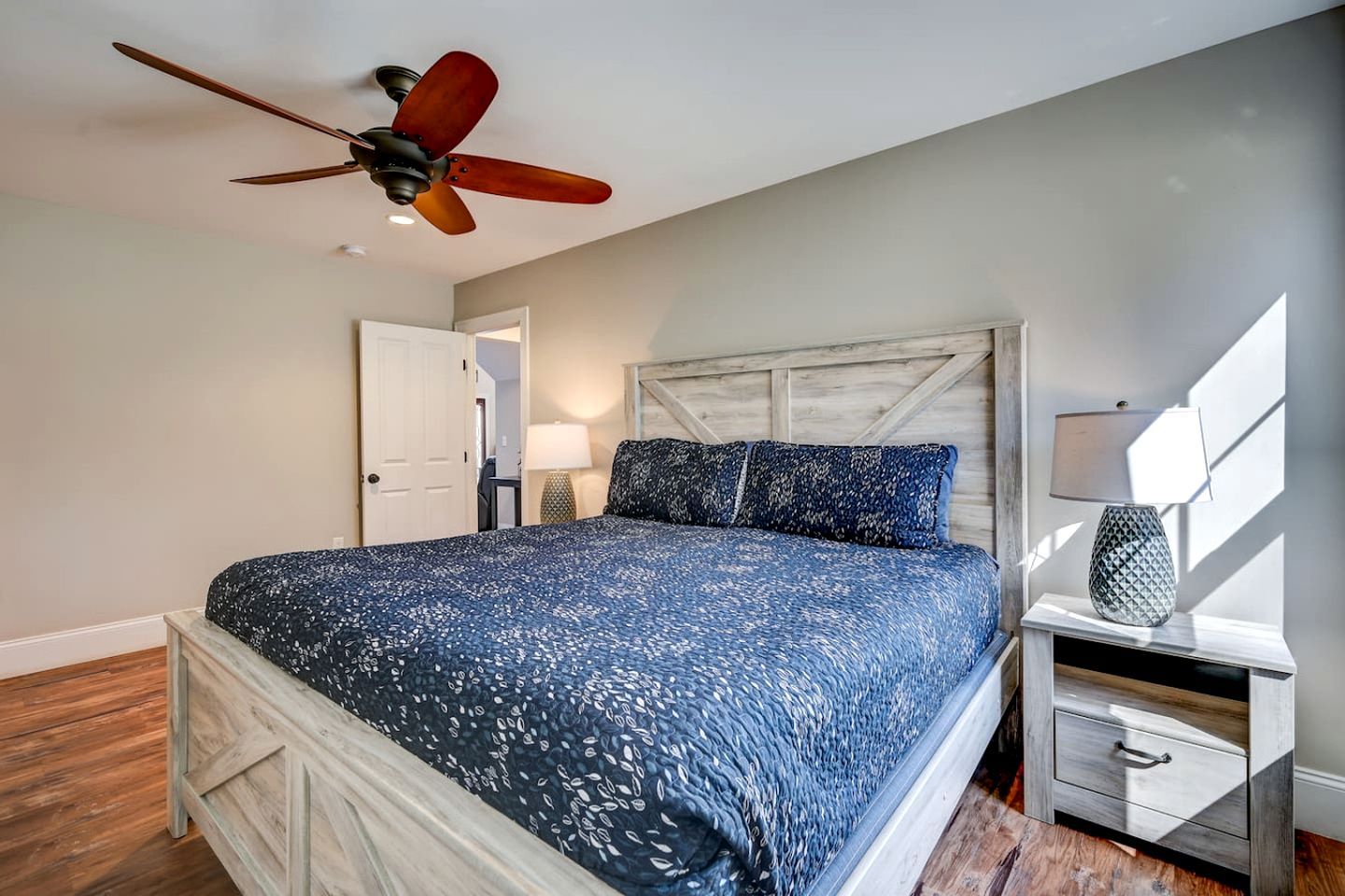 A bedroom with a bed, a nightstand, a lamp, a ceiling fan, and a door.
