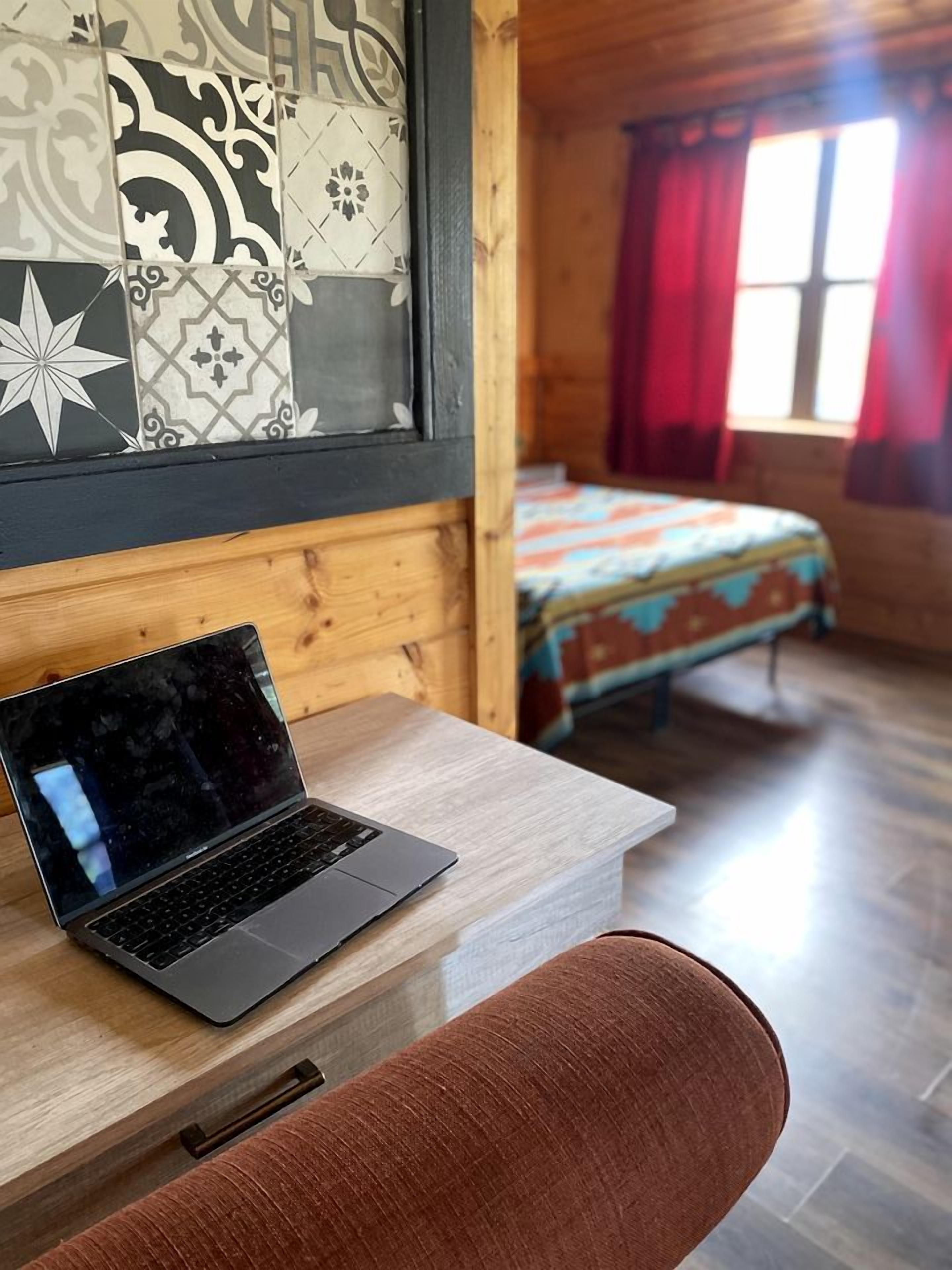Ein Interieur einer Holzhütte mit einem Laptop auf einem Schreibtisch, einem Bett mit bunter Bettdecke und einem Fenster mit roten Vorhängen.