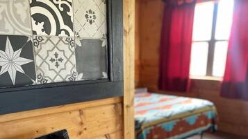 Ein Interieur einer Holzhütte mit einem Laptop auf einem Schreibtisch, einem Bett mit bunter Bettdecke und einem Fenster mit roten Vorhängen.