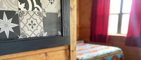 A wooden cabin interior with a laptop on a desk, a bed with a colorful bedspread, and a window with red curtains.