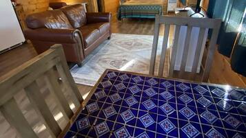 Interior acogedor de una cabaña con techo de madera, una cama con colcha estampada, un sofá de cuero y una mesa de comedor sobre piso de azulejos azules.