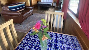 A wooden cabin interior with a bed, a brown leather couch, a dining table with a blue patterned tablecloth, and a window with red curtains.