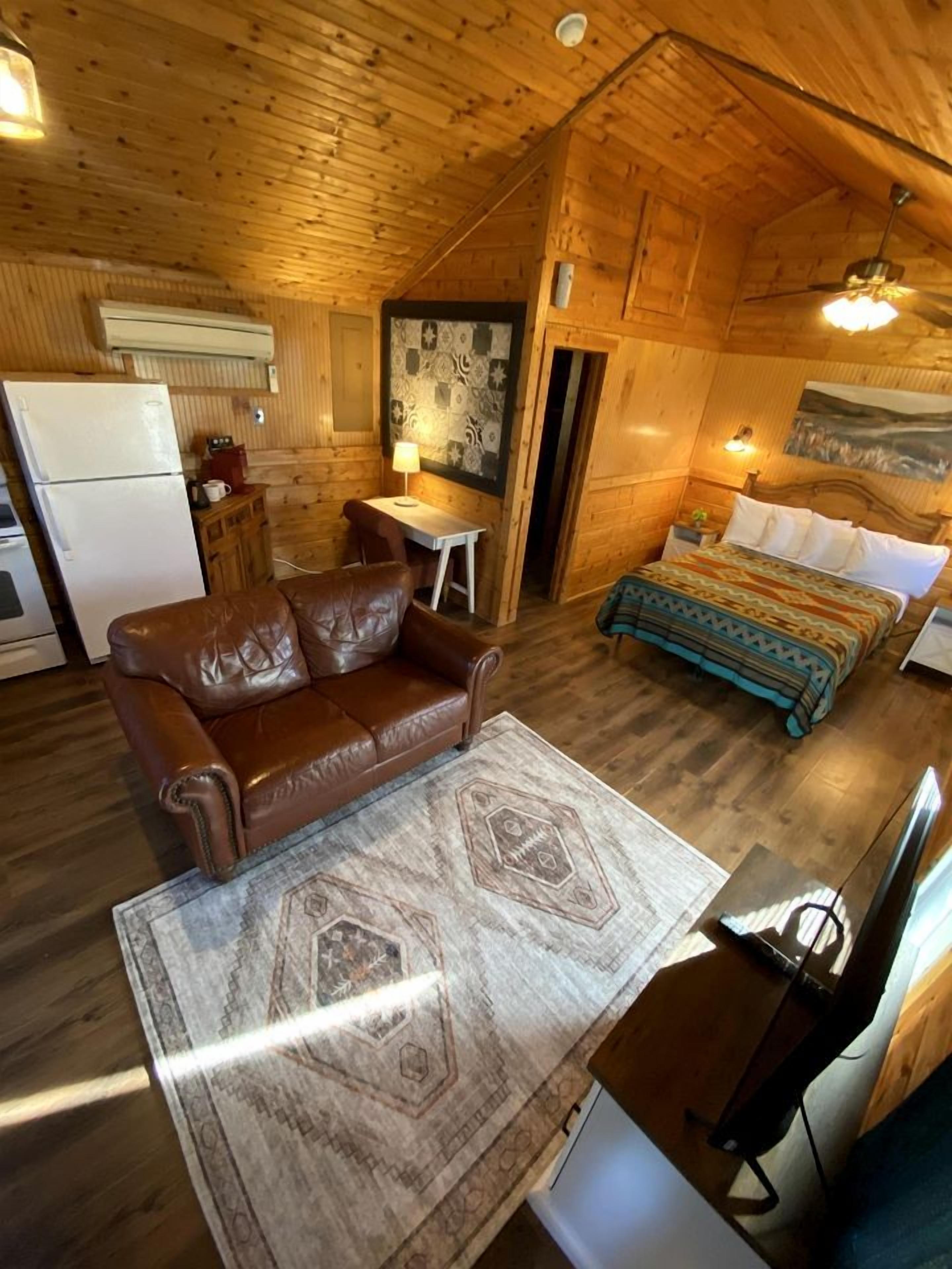 A wooden cabin interior with a sofa, a bed with patterned bedding, a small table, and a refrigerator.