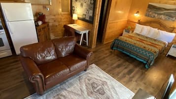A wooden cabin interior with a sofa, a bed with patterned bedding, a small table, and a refrigerator.