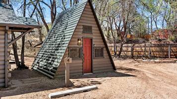 A small, single-story log cabin with a red door and a sloped roof, surrounded by trees and a dirt path.