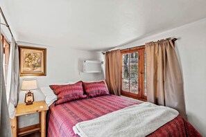 A bedroom with a bed, a bedside table with a lamp, a picture on the wall, and a window with curtains. - Charming A-Frame Cabins with Scenic Views, Nestled Near Garden of the Gods in Colorado Springs, CO (Colorado Springs)