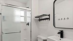 Kylpyhuone, jossa on valkoinen wc-istuin, mustalla hanalla varustettu pesuallas, pyyhekoukku ja lasinen suihkuovi.