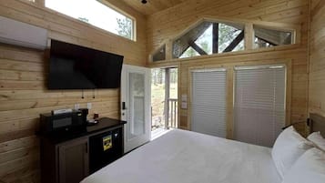 A wooden cabin room with a bed, a flat-screen TV, a desk with a computer, and a sliding door to the outside.