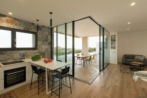 Una cucina moderna con una grande isola, un'area pranzo con tavolo e sedie, e un soggiorno con divano e vista sull'esterno.