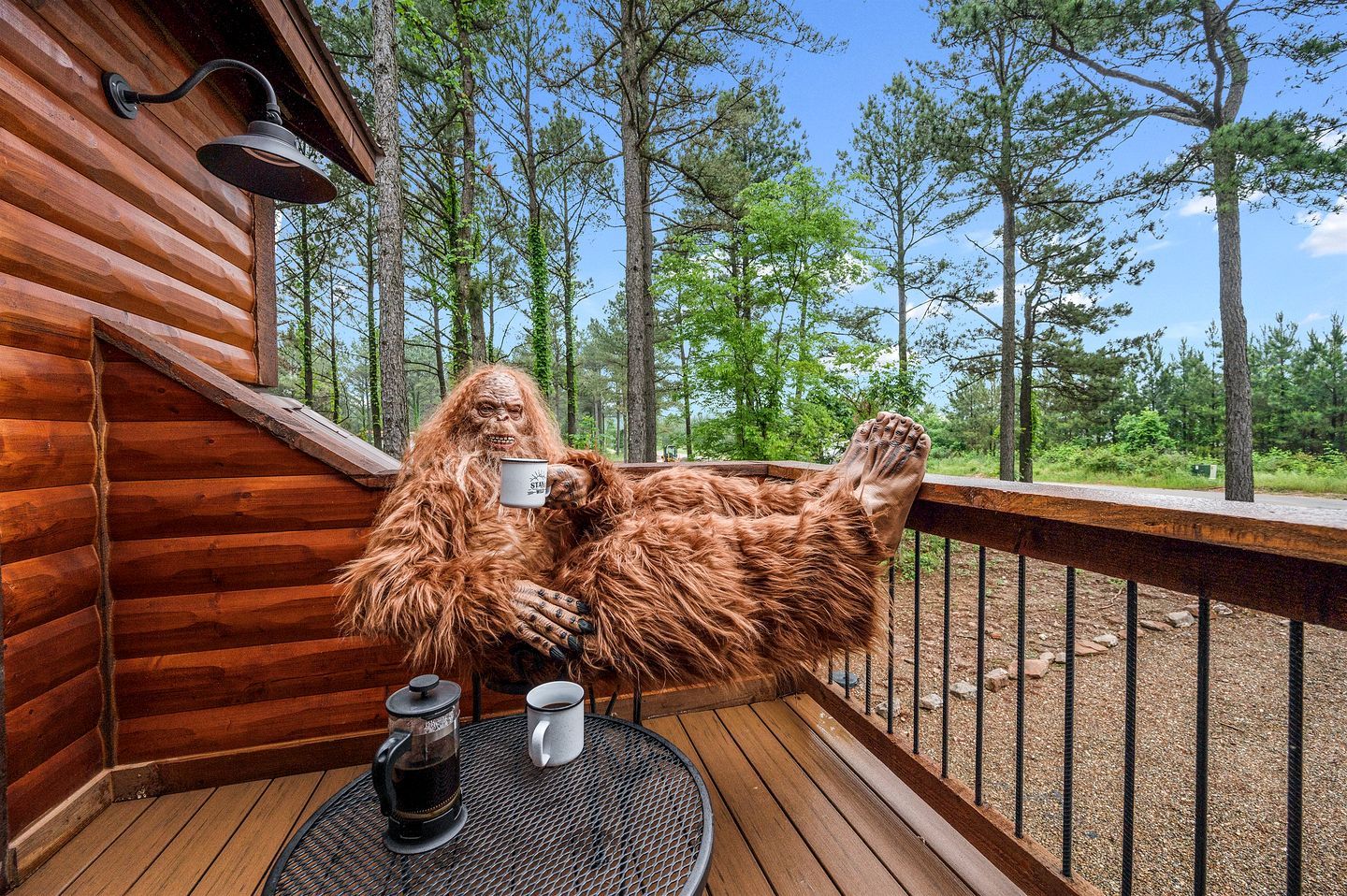 Een houten blokhut met terras, een tafel met koffiepot en mok, en een persoon in een behaarde kostuum.