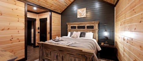 A wooden-paneled bedroom with a large bed, a ceiling fan, and a nightstand with a lamp.