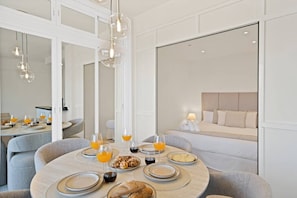 Une chambre d’hôtel moderne dotée d’un coin repas avec une table ronde dressée pour le petit-déjeuner, un lit avec une tête de lit et un grand miroir qui reflète la pièce.