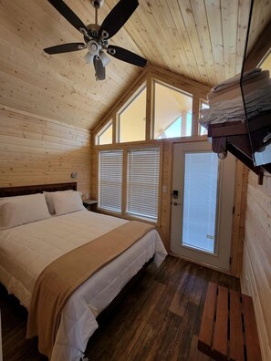 A bedroom with a bed, a ceiling fan, wooden walls, a window, and a door. - Modern Tiny Home Retreat with Stunning Stargazing Views in Williams, Arizona (Williams)