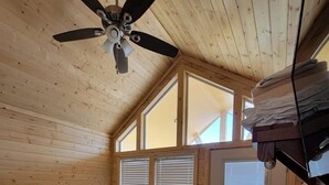 Quarto com cama, ventilador de teto, paredes de madeira, janela e porta.