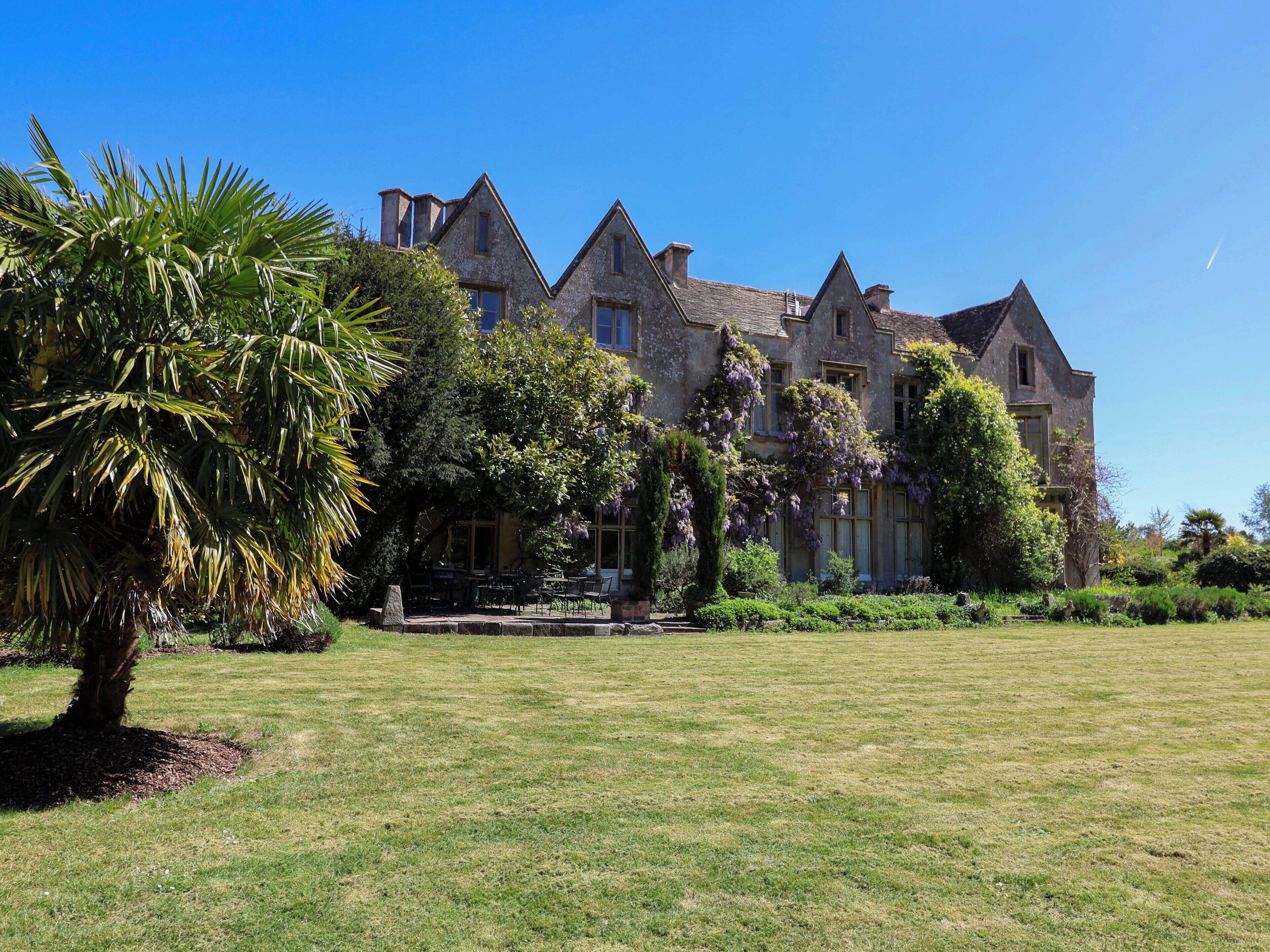 A large, historic building with multiple chimneys, surrounded by a well-maintained lawn and various trees.