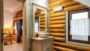 Ein Badezimmer im Stil eines Holzblockhauses mit einer Holzwand, einem großen Spiegel, einem Holzkabinent und einem Waschbecken mit dekorativer Schale. Ein Fenster mit Vorhang vervollständigt das Bild.