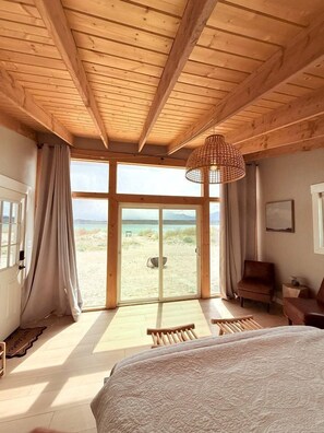 A room with a bed, a chair, a hanging light fixture, and a view of the beach through large glass doors.