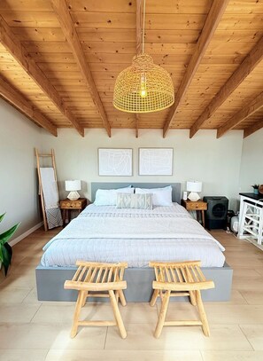 A bedroom with a wooden ceiling, a bed with a grey headboard, two bedside tables with lamps, a small desk, and a chair.