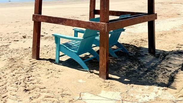 Une structure en bois installée sur la plage, dotée d’un toit de chaume et entourée de chaises turquoise.
