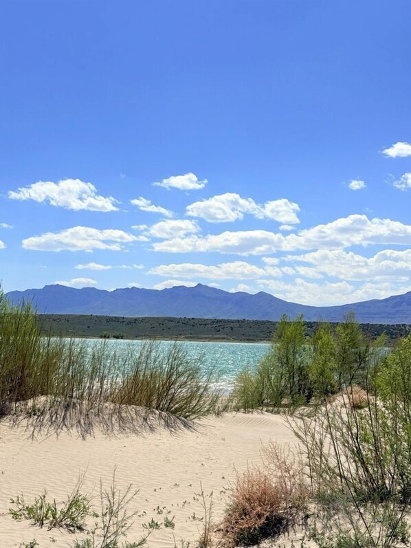 Una playa con agua cristalina, dunas de arena y montañas al fondo.