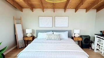 A bedroom with a wooden ceiling, a bed with a grey headboard, two bedside tables with lamps, a small desk, and a chair.