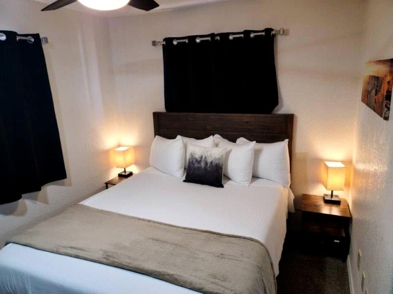 A hotel room with a bed, two bedside tables, a ceiling fan, and black curtains.