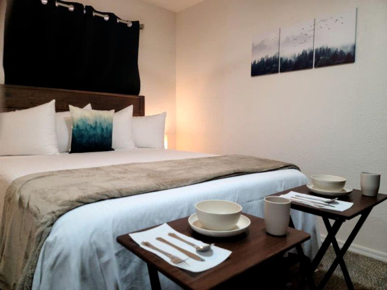 A hotel room with a bed, a bedside table with a bowl and utensils, a wall-mounted painting, and a window with curtains.