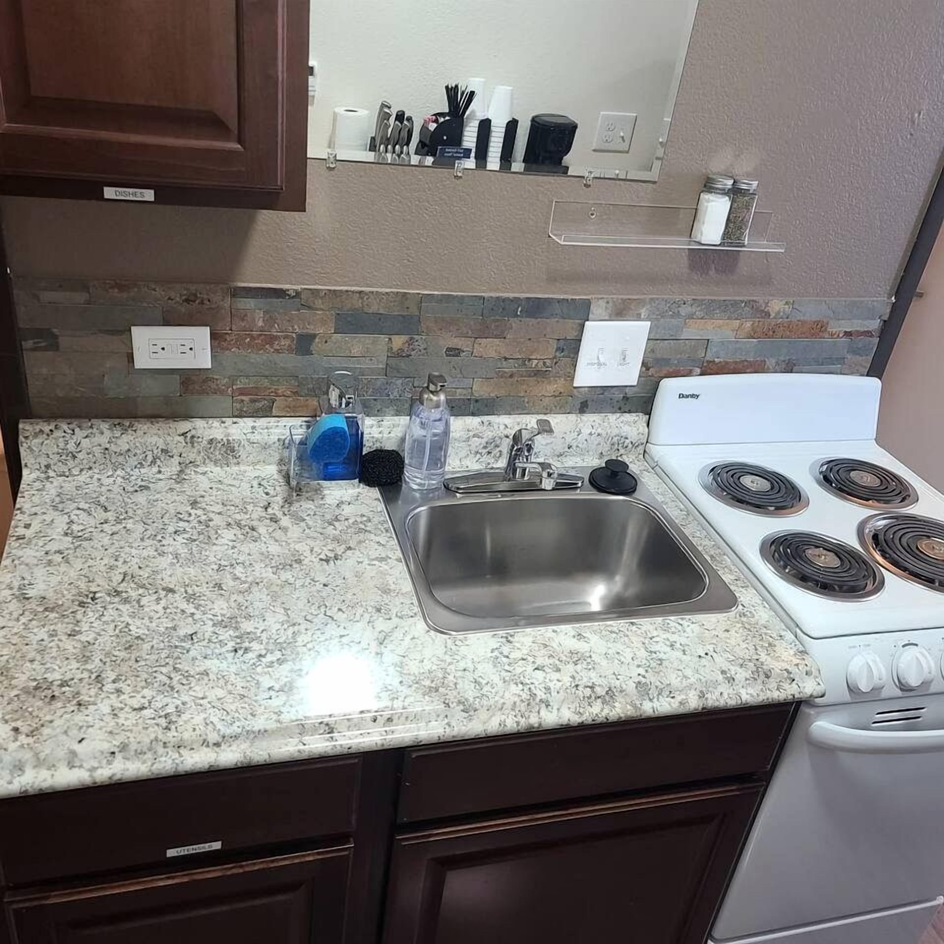 A kitchen with granite countertops, a stainless steel sink, a white Danby stove, and a stone backsplash.