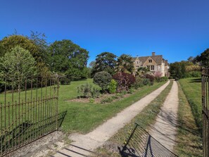 Ein Weg führt zu einem großen, historischen Haus, das von üppigem Grün und einem Metallzaun umgeben ist.
