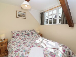 A bedroom with a bed, a nightstand, a lamp, a window with curtains, and a picture on the wall. - The Old Stables (Gloucester)