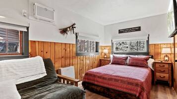 A bedroom with a bed, a chair, a bedside table, a lamp, a wall-mounted TV, and a window with a view of trees.