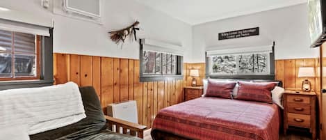 Une chambre à coucher comprenant un lit, une chaise, une table de chevet, une lampe, un téléviseur fixé au mur et une fenêtre donnant sur des arbres.