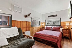 Ein Schlafzimmer mit Bett, Sessel, Nachttisch, Lampe, einem an der Wand montierten Fernseher und einem Fenster mit Blick auf Bäume.