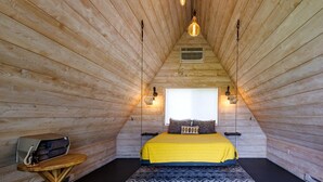 Ein gemütliches Schlafzimmer im Dachgeschoss mit schrägen Wänden, einem Bett mit gelber Bettdecke, einem kleinen runden Tisch mit Lampe und einem Fenster mit Blick.