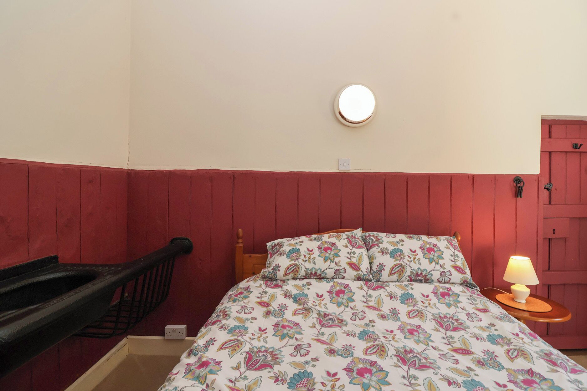 Une chambre à coucher avec un lit, une table de chevet avec une lampe, une chaise en bois, une porte rouge et un luminaire fixé au mur.