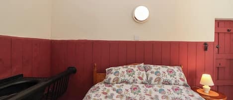Une chambre à coucher avec un lit, une table de chevet avec une lampe, une chaise en bois, une porte rouge et un luminaire fixé au mur.
