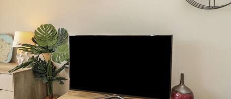 A modern living room with a flat-screen TV on a wooden table, a wall clock, a potted plant, and a decorative vase.