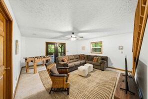 Ein Wohnzimmer mit Ecksofa, einem Kickertisch aus Holz, Deckenventilator und Fernseher.