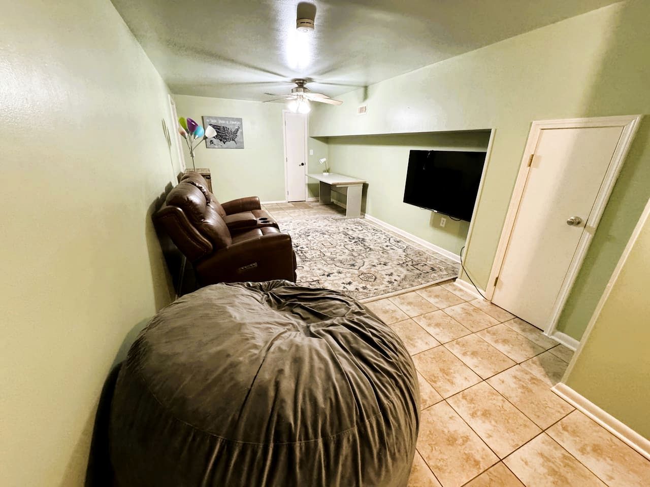 A room with a ceiling fan, a brown leather chair, a large bean bag, a flat-screen TV, and a door.